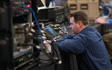 Brown haired man wearing a blue plaid shirt looking at an equipment screen in a server room